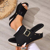 Black open-toe sandals with a gold buckle worn by a person on a wooden floor.