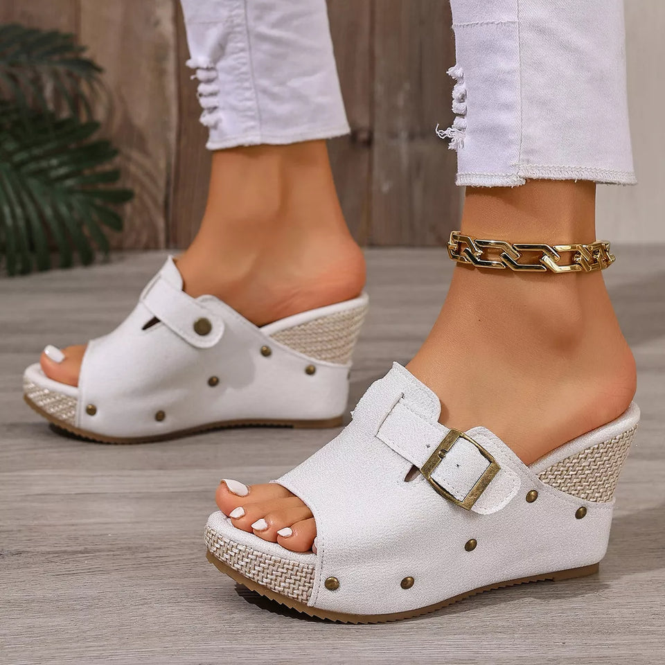 White wedge sandals with gold buckles worn by a person on a wooden floor.